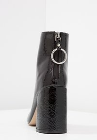 Schwarze Lackleder-Ankle Boots mit Absatz, ausgestattet mit einem Reißverschluss und einem runden Metall-Detail am Reißverschlusszug.