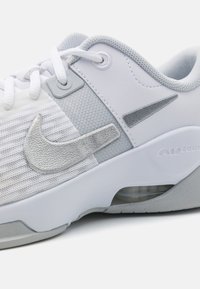 Scarpa sportiva bianca e grigia chiara con materiale in rete, accenti testurizzati e un'unità d'aria visibile. Presenta un logo Nike in argento e dettagli cuciti.