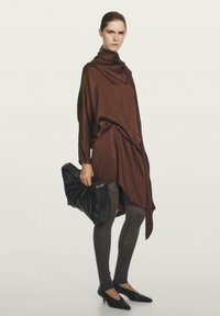 Bruine gedrapeerde jurk met een zachte textuur, gecombineerd met grijze legging en zwarte puntige ballerina's. Model houdt een zwart pouch-stijl tas vast.