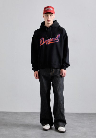 Tânăr îmbrăcat cu un hanorac negru Dsquared2, blugi negri cu croială largă, adidași albi și o șapcă roșie Dsquared2, stând pe un pardoseală gri.