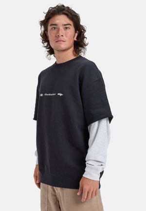 Sweatshirt noir surdimensionné à manches courtes, arborant un logo blanc sur la poitrine. Superposé sur une chemise à manches longues gris clair.