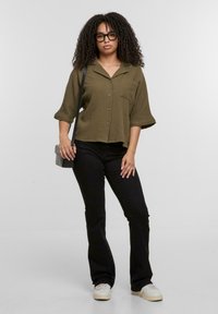 Camicia verde oliva con maniche corte e bottoni, con tasca frontale; jeans neri a campana; sneaker bianche; borsa a tracolla; capelli ricci.