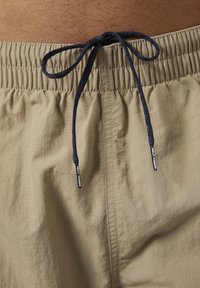 Pantalones cortos de baño beige con cintura elástica, que cuentan con un cordón de marinero. Textura suave y detalles mínimos.