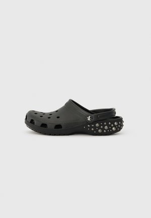 Sabot en caoutchouc noir avec des trous de ventilation, une semelle robuste et des clous décoratifs en argent sur le talon. Sangle arrière réglable pour un maintien sûr.