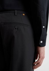 Schwarze maßgeschneiderte Hosen mit einer Gesäßtasche, die mit einem Knopfverschluss versehen ist. Die Hemdmanschetten sind mit Knöpfen gestaltet, beide in einem eleganten Design.