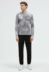Graues Langarmshirt mit einem Mischdruck aus Typografie, kombiniert mit schwarzen Hosen und weißen Sneakern. Sanfte Stofftextur und lässiges Design.