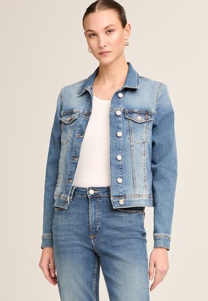 Frau trägt eine hellblaue Jeansjacke über einem weißen Top und passende blaue Jeans, steht vor einem schlichten beigefarbenen Hintergrund.