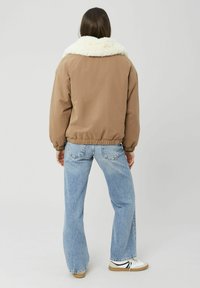 Veste aviateur marron avec un col en fausse fourrure blanche, des poignets et un ourlet élastiques, associée à un jean évasé bleu clair et des baskets blanches.