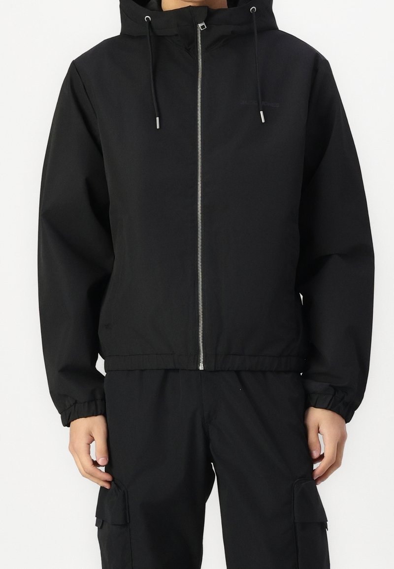 Personne portant une veste à capuche noire avec fermeture éclair, avec des cordons de serrage et un pantalon cargo noir assorti, sur fond uni.