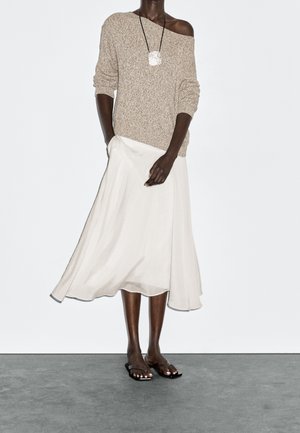 Osoba o ciemnej karnacji ubrana w beżowy sweter z odkrytymi ramionami, białą, zwiewną spódnicę midi, czarne sandały oraz srebrny naszyjnik z zawieszką.