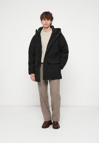 Casaco acolchoado preto com capuz, com fecho de correr e fecho de pressão, dois bolsos frontais, usado sobre um suéter leve de malha com tranças e calças bege.