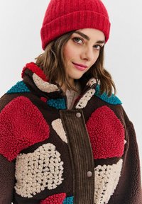 Mladá žena s červenou pletenou čiapkou a farebnou patchwork sherpa bundou s textúrovanými sekciami v červenej, krémovej, modrej a hnedej farbe.