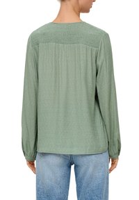 Lange mouw, lichtgroen shirt van een gestructureerde stof met smockdetails op de achterkant, gecombineerd met blauwe denim jeans.