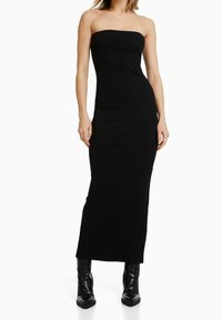 Robe maxi noire sans bretelles avec une silhouette ajustée, présentant une texture lisse et un décolleté droit. Portée avec des bottines noires.