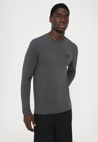 Young man wearing a fitted dark gray long-sleeve shirt with a small "EA7" logo on the chest, standing against a plain light background.