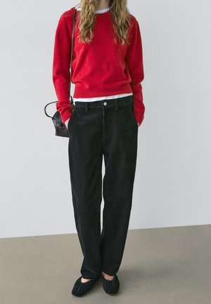 Femme portant un pull rouge par-dessus une chemise blanche, un pantalon noir, des chaussures noires, et portant un petit sac à bandoulière noir, debout les mains dans les poches.