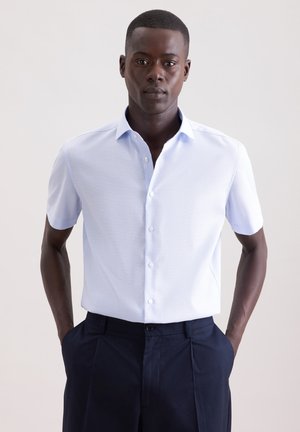 Mężczyzna w jasnoniebieskiej, krótkorękawowej koszuli z guzikami, włożonej do ciemnogranatowych spodni, stoi z rękami w kieszeniach na jednolitym tle.