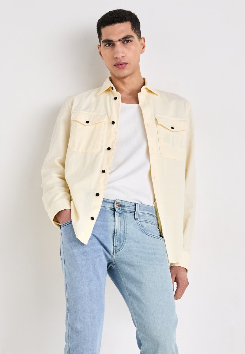 Lichtgele button-up shirt met twee borstzakken en zwarte knopen, gecombineerd met lichtblauwe recht-toe-rechtdoor jeans en een witte tanktop.