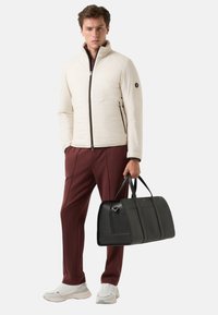 Helles beige gepolstertes Jacket mit Reißverschluss, schwarzen Akzenten, kastanienbraunen Hosen und einer schwarzen Ledertasche. Weiße und graue Sportschuhe.