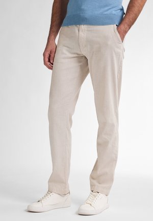 Mężczyzna w beżowych lnianych spodniach, białych trampkach i jasnoniebieskim swetrze, stojący z jedną ręką w kieszeni.