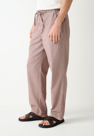 Uomo che indossa pantaloni larghi marrone chiaro con coulisse e sandali intrecciati marrone scuro, in piedi davanti a uno sfondo bianco semplice.
