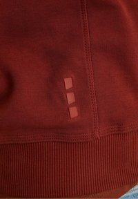 Sweat-shirt rouge en tissu texturé, ourlet côtelé, avec trois accents carrés en relief près de la couture. Design simple aux lignes épurées.