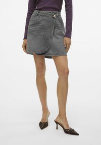 Falda envolvente de denim gris con una cintura alta, que presenta un cierre de botón, detalles de costura y un dobladillo ligeramente irregular. Complementada con zapatos de tacón marrones.