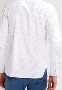 Bílá košile s malými černými puntíky, manžetami na knoflíky a zakřiveným lemem. Pohled zezadu ukazuje hladkou texturu látky a minimalistické designové detaily.