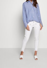 Niebiesko-biała bluzka w paski z długimi rękawami, zestawiona z białymi, obcisłymi dżinsami z postrzępionymi mankietami i białymi klapkami.