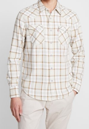 Mies, jolla on beige-valkoinen ruudullinen nappipaita, jossa kaksi rintataskua, ja beiget housut, kädet rentoina sivuilla.