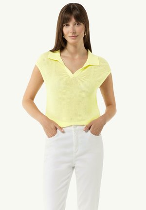 Dzianinowy sweter polo w pastelowym żółtym kolorze z dekoltem w serek, krótkimi rękawami i teksturowanym splotem. Zestawiony z białymi spodniami, ręce spoczywają w kieszeniach.