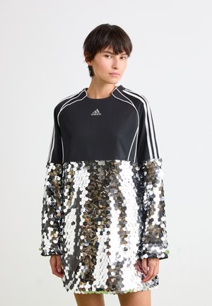 Woman wearing a black Adidas top with white stripes and large silver sequined skirt against a plain white background.