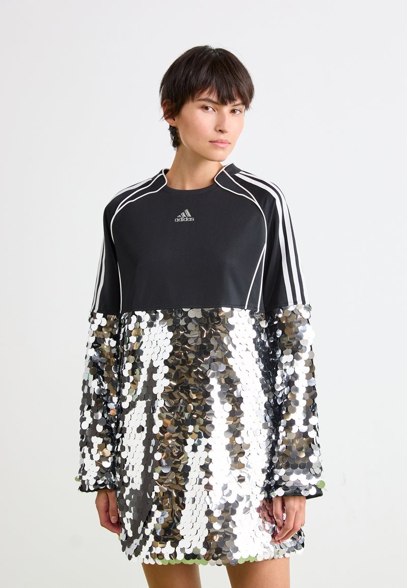 Sieviete ģērbusies melnā Adidas topā ar baltām svītrām un lielā sudraba mirdzošā svārku fona priekšā.