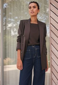 Blazer marrone scuro con bottoni doppiato sopra una maglia marrone, abbinato a jeans denim a gamba larga con cuciture a contrasto e tasche.