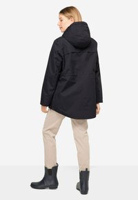 Chaqueta negra impermeable con capucha, corte relajado y cintura elástica. Combinada con pantalones beige y botas negras de tobillo.