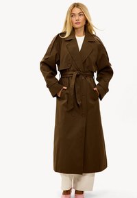 Femme aux longs cheveux blonds, portant un trench-coat marron foncé ceinturé, un pantalon blanc et des chaussures roses, se tenant les mains dans les poches.