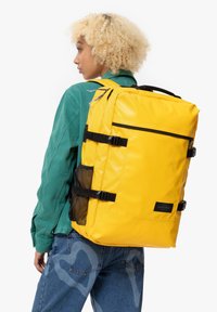 Gul ryggsäck med slät, glänsande yta; har svarta detaljer, justerbara remmar och en mesh-sidoficka. Bärs på ena axeln.