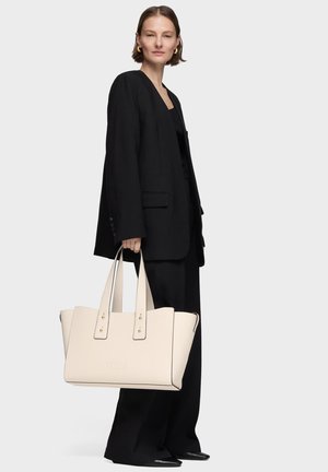 Mujer con traje negro sosteniendo un gran bolso tote beige con asas dobles, de pie frente a un fondo liso y claro.