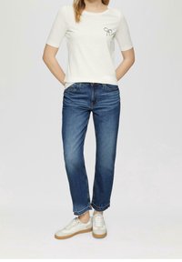 Camiseta blanca de manga corta con un motivo de lazo en el pecho, combinada con jeans azul de pierna recta y zapatillas blancas con suela marrón.