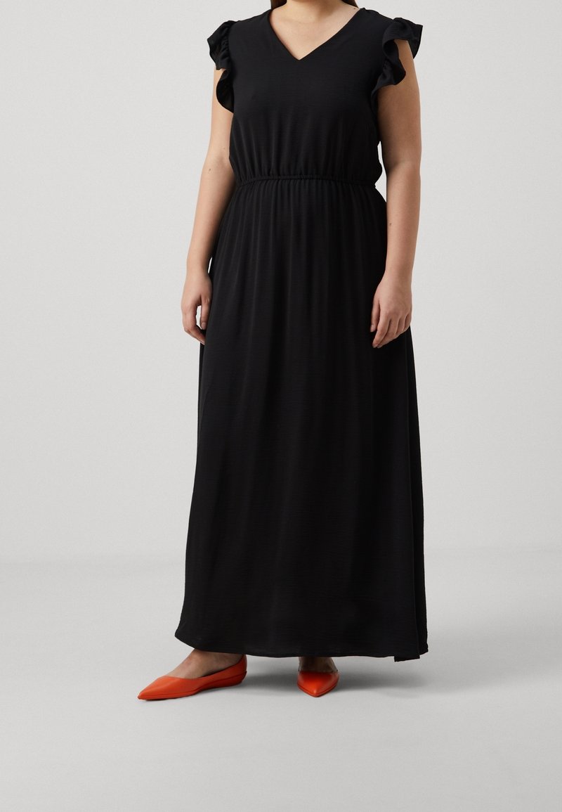 Kvinna i en svart ärmlös maxiklänning med volangärmar och klarröda spetsiga ballerina-skor, stående mot en enfärgad bakgrund.