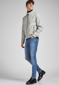 Jachetă bomber gri deschis cu o textură netedă, guler negru și fermoare, asortată cu blugi albastri slim-fit și cizme negre cu șireturi.