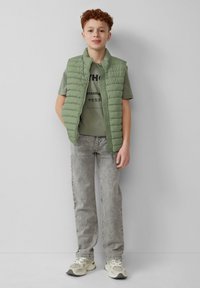Hellgrüne gepolsterte Weste über einem grauen Graphic-T-Shirt, kombiniert mit hellgrauen, ausgebleichten Jeans und beigen Sneakers, vor einem neutralen Hintergrund.