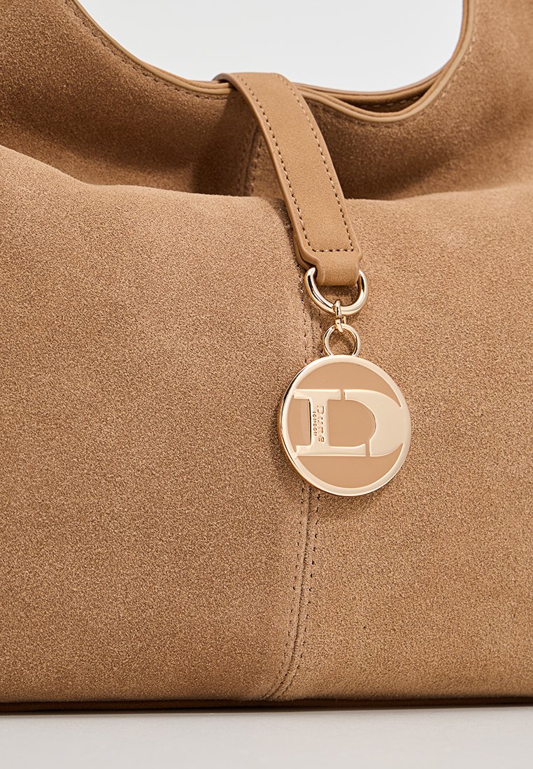 Nahaufnahme einer hellbraunen Wildlederhandtasche mit passendem Schulterriemen und einem rundem goldfarbenem Emblem-Anhänger, der vom Riemen hängt.