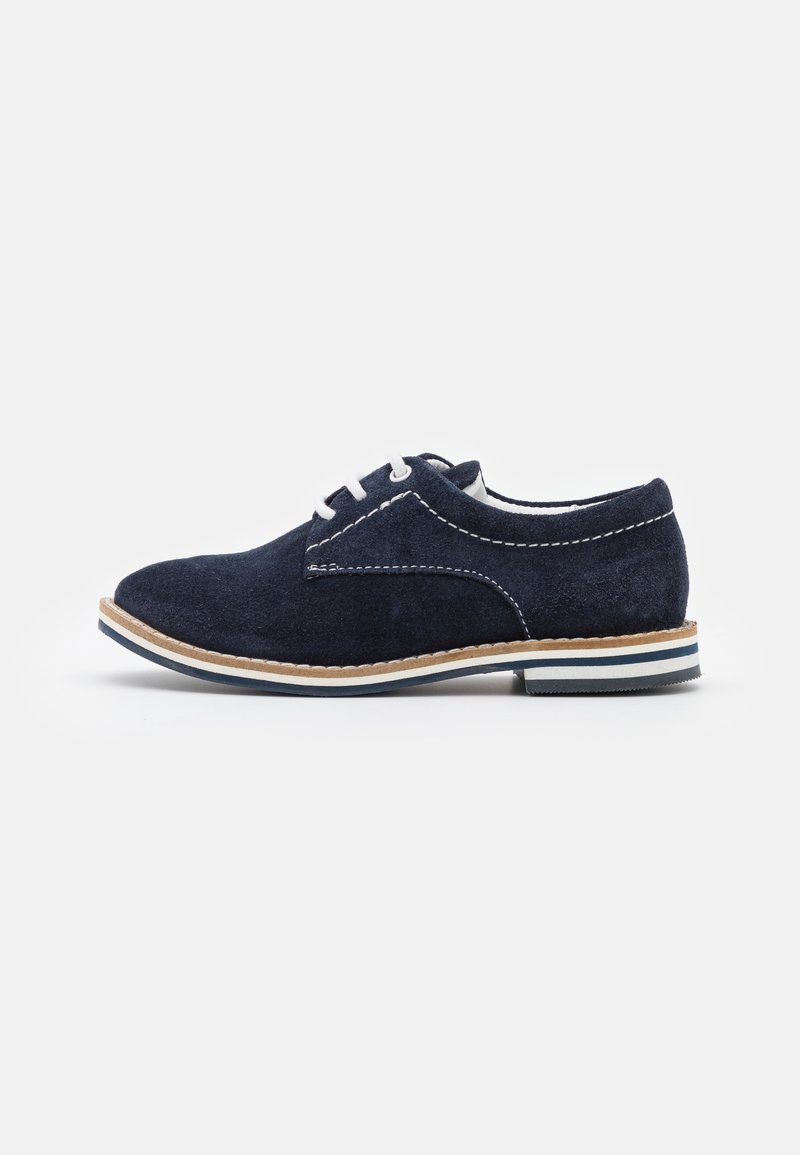 Scarpa stringata in camoscio blu navy con cuciture bianche, lacci bianchi e suola caratterizzata da strisce beige e blu navy su sfondo bianco.
