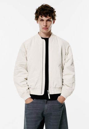 Hellbeige Bomberjacke mit Reißverschluss, gerippten Bündchen und Seitentaschen, kombiniert mit dunkelgrauen Hosen in einem minimalistischen Umfeld.