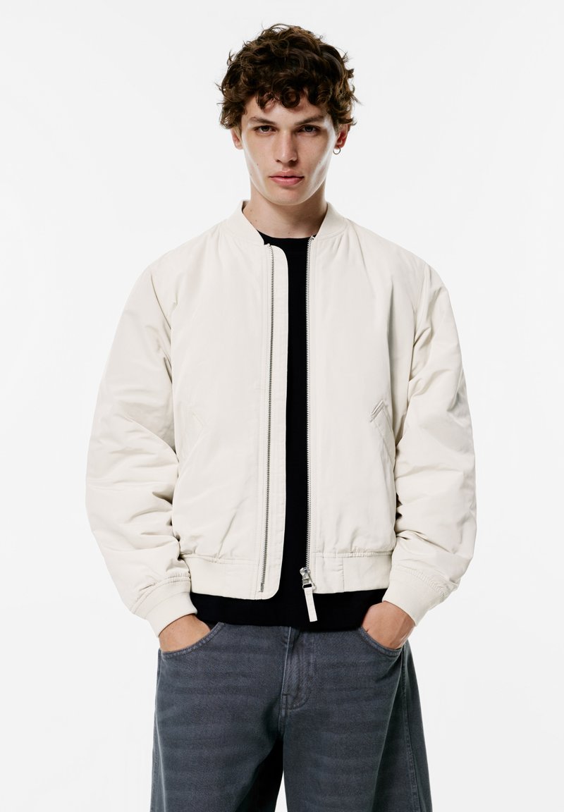 Light beige bomber jacket with a zip closure, ribbed cuffs, and side pockets, paired with dark gray pants in a minimalistic setting.