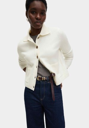 Frau trägt eine weiße, geknöpfte Strickjacke über einem grauen Oberteil, dunkelblaue Jeans und einen braunen Gürtel mit goldener Schnalle.
