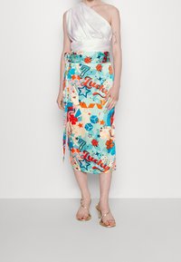 Nainen, joka on pukeutunut valkoiseen olkaimeton toppiin ja värikkääseen midihameeseen, jossa on onnekkaita symboleja, kuten leppäkerttuja, noppia, tähtiä, apiloita ja kultaisia hihnatossuja.