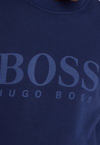 Marinblå sweatshirt gjord av texturerad tyg med stora, ljusblå bokstäver av "BOSS" och "HUGO BOSS" i en modern stil.