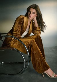 Traje de terciopelo marrón con estampados florales, combinado con una blusa blanca y sandalias de tacón beige. Sentada en una silla plateada frente a un fondo degradado.